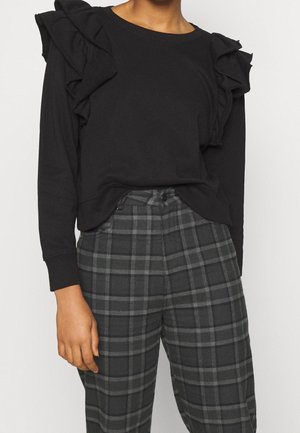 Kobieta ubrana w czarną bluzkę z długim rękawem z falbankami na ramionach oraz wysokim stanem ciemnoszare spodnie w kratę na jednolitym tle.