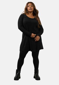 Abito lungo nero a coste con scollo rotondo e orlo sfilacciato, abbinato a leggings neri e stivali neri con tacco largo.