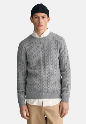 Gray cable-knit sweater in soft fabric, ribbed cuffs and hem, layered over a white collared shirt. Black beanie completes the look.