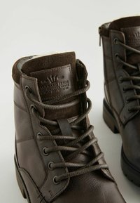 Botas de cuero marrón con acentos de ante, superficies texturizadas, cordones redondos y un cuello acolchado. Etiqueta de marca en el lado.
