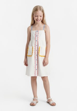 Young girl smiling, wearing white sleeveless dress with blue straps, pink and yellow scalloped trim, front buttons, and side pockets, standing barefoot.