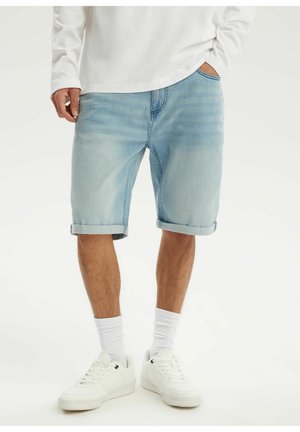 Mężczyzna noszący jasnoniebieskie, podwinięte szorty jeansowe, białą koszulę z długim rękawem, białe skarpetki oraz białe sneakersy, stoi z rękami w kieszeniach.