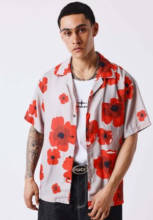 Jeune homme aux cheveux courts et noirs portant une chemise beige avec de grands motifs floraux rouges et noirs, un T-shirt blanc imprimé et un jean foncé.