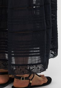 Robe maxi noire en dentelle transparente avec un motif à rayures horizontales et un ourlet en dentelle complexe. Portée avec des sandales noires ornées de détails en or.
