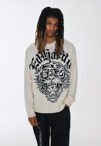 Maglione a maglia crema con una grande grafica di una tigre nera e un testo audace, con polsini e orlo a costine. Indossato con pantaloni larghi scuri.
