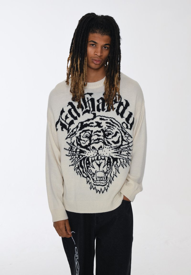 Maglione a maglia crema con una grande grafica di una tigre nera e un testo audace, con polsini e orlo a costine. Indossato con pantaloni larghi scuri.