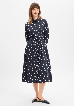 Femme portant une robe midi bleu marine à manches longues à pois, avec une ceinture à la taille et des ballerines slingback noires à bout pointu, debout sur un fond blanc.