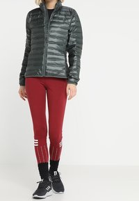 Žena v tmavě šedé péřové bundě, bordó legínách s bílými pruhy u lýtek, černých ponožkách a černých sportovních botách.