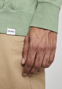 Felpa verde chiaro con polsini e orlo a coste, con un'etichetta bianca riportante il testo "PULL&BEAR", indossata sopra un tessuto beige. Dettagli delle mani in vista.