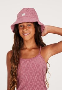 Sombrero de cubo de algodón de color rosa claro con una superficie texturizada. Lleva puesto una chica con cabello largo, combinado con una blusa de tirantes texturizada a juego.