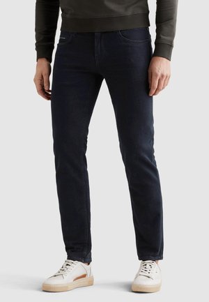 Mand iført mørkeblå slim-fit jeans, hvide sneakers og en mørkeolivengrøn sweatshirt, stående mod en enkel lys baggrund.