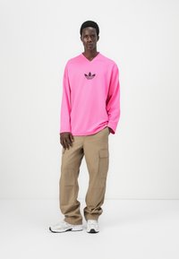 Långärmad rosa träningsskjorta med en svart logotyp, i kombination med khaki cargobyxor. Modellen bär vita sneakers. Enfärgad bakgrund.