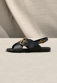 Μαύρο δερμάτινο σανδάλι με φαρδιές σταυρωτές λουριά και μεγάλες αγκράφες σε χρυσό τόνο, επίπεδη σόλα και ρυθμιζόμενο λουράκι στον αστράγαλο σε ουδέτερο φόντο.