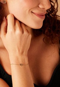 Gouden kettingarmband met een hartvormige hanger met decoratieve accenten. Het ontwerp omvat een delicate ketting en een gestructureerd hart.