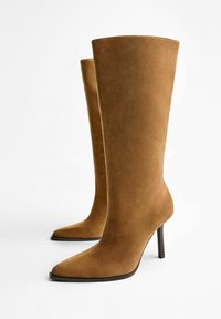 Bottes en daim marron jusqu'au genou avec un bout pointu et un talon aiguille. Texture lisse avec un design minimaliste et sans quincaillerie visible.