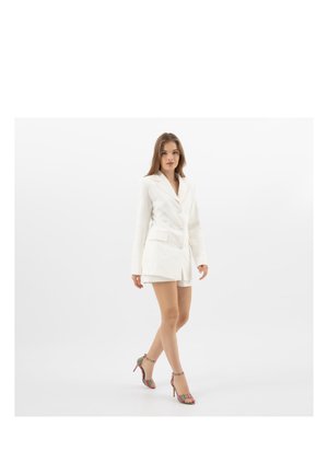 Frau geht in weißem Blazer-Kleid mit Knöpfen, kombiniert mit bunten, gestreiften Sandalen mit hohen Absätzen auf weißem Hintergrund.