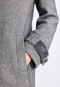 Grå hundtandsmönstrad kappa med texturerat tyg, fickor med påsydda lappar och en märkesetikett på ärmsluten med knappstängning.