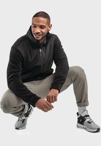 Schwarze Fleece- und Softshell-Jacke, Reißverschluss vorne, graue elastische Bundhose, graue Sneakers mit schwarzen Akzenten und strukturiertem Obermaterial.