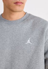 Sudadera gris de algodón, con cuello redondo y un logo Jumpman bordado en blanco en el lado izquierdo del pecho. Textura suave.