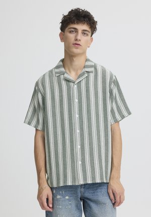 Giovane uomo con capelli ricci che indossa una camicia a maniche corte con bottoni a righe verticali verdi e bianche e jeans blu consumati.