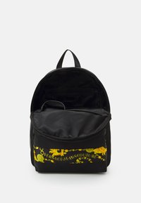 Sac à dos noir avec une texture lisse, arborant un motif doré coloré et un logo sur le devant. Comprend un intérieur spacieux et une poche zippée.