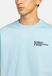 Camiseta de algodón azul claro con cuello redondo y mangas cortas, con un logo impreso en negro en el lado izquierdo del pecho.