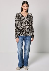 Blusa beige a maniche lunghe con stampa leopardata nera, scollatura a V e dettaglio arricciato alle spalle, abbinata a jeans svasati azzurri chiari e sneakers.