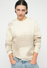 Kremasti pulover s svijetlosmešavim apstraktnim uzorcima, rebrasti okrugli izrez i manžetne, mekana tekstura, uparen s svijetloplavi trapericama.