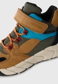 Zapato de senderismo marrón y verde con un acento azul, cordones naranjas, cuero texturizado y una suela de goma blanca con un patrón en zigzag.