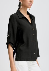 Camicia nera con bottoni, colletto e maniche a tre quarti, realizzata in un tessuto texturizzato e con spacchi laterali, completata da bottoni dorati.