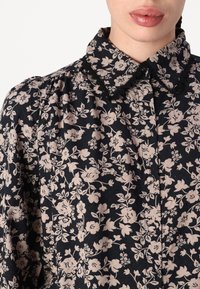 Camisa negra con estampado floral y flores rosa claro, con cuello adornado con encaje y un corte relajado. Material suave con un brillo sutil y tejido texturizado.