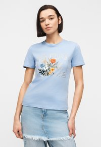 Femme portant un t-shirt bleu clair à imprimé floral avec le texte « LES FLEURS », associé à une jupe en denim clair effilochée par-dessus un jean.