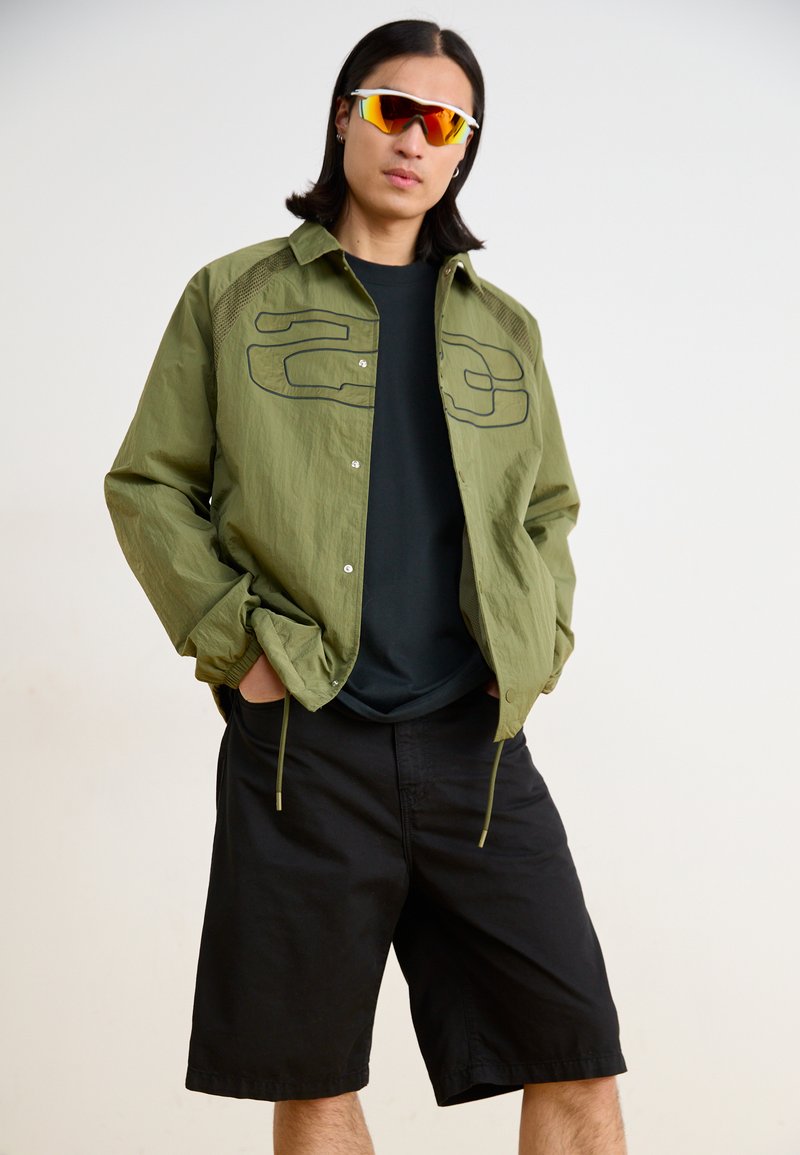 Lightweight olive green jacket with black accents, short sleeves, and two large front pockets, paired with black shorts and sporty sunglasses.