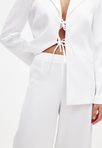 Valge kahest osast koosnev kostüüm, millel on tekstuuriga jakk, millel on paelaga sulgemine, ja laiad püksid. Mõlemal osal on sile, kerge kangas.