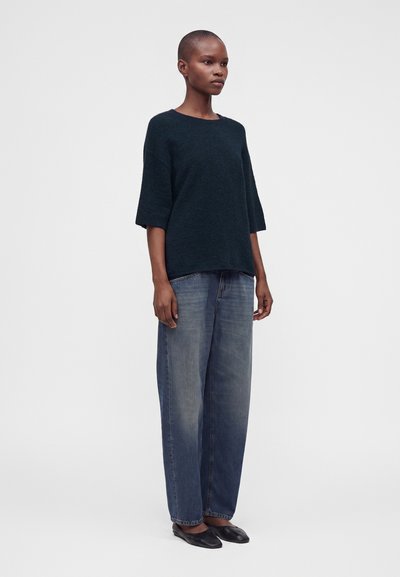 Personne debout portant un pull sombre et ample, un jean large bleu délavé et des chaussures plates noires devant un fond clair uni.
