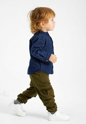 Barn i en blå ternet skjorte med knap og olivengrønne cargo-bukser, parret med hvide sneakers. Håret er stylet i løse krøller.