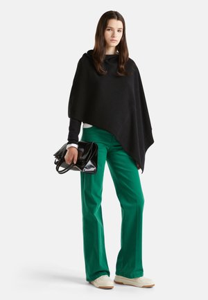 Poncho noir avec capuche, pantalon évasé vert, et sac à main noir brillant. Baskets blanches mi-mollet avec des accents beiges.