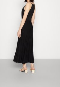 Femme portant une robe midi plissée noire sans manches et des sandales à talons beiges, se tenant devant un fond blanc uni.