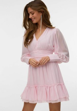 Femme aux longs cheveux bruns portant une robe plissée rose clair avec des manches bouffantes et un décolleté en V, souriante et regardant vers le bas.