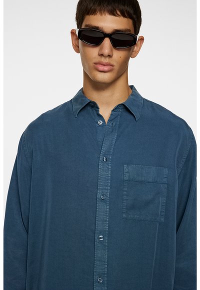 Blaues Hemd mit Knöpfen aus leichtem Stoff, ausgestattet mit einer Brusttasche, klassischem Kragen und entspannter Passform. Das Modell trägt schwarze Sonnenbrillen.