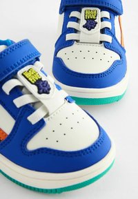 Baskets bleu et blanc avec une boîte à orteils perforée, un accent orange contrastant, une sangle Velcro, des lacets et une semelle en caoutchouc verte.