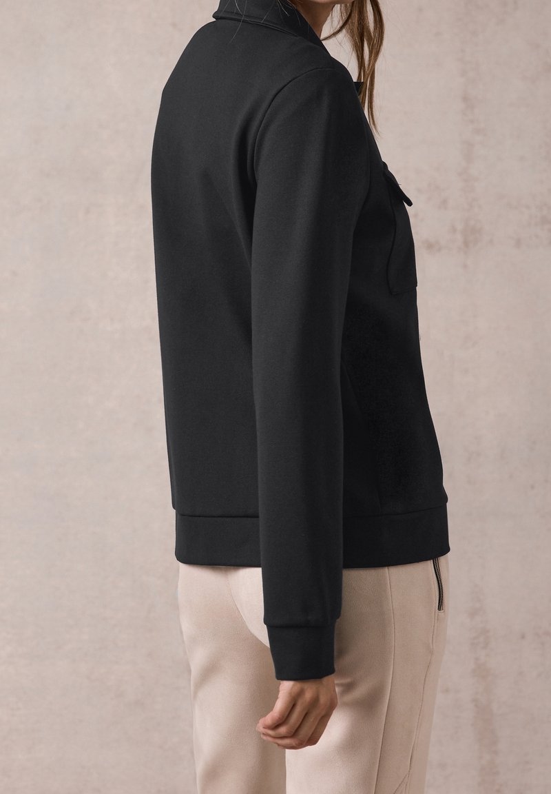Femme vue de profil portant une veste noire à manches longues et un pantalon beige clair avec une poche latérale zippée sur fond neutre.
