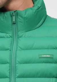 Geacă puffer verde cu guler înalt, cu fermoar frontal și panouri cusute. Pachet cu logo Calvin Klein pe partea de jos.