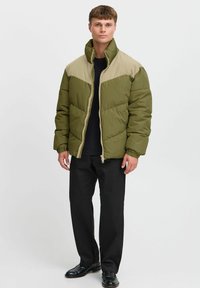 Chaqueta acolchada verde oliva con hombros beige, costuras horizontales, cuello alto, cremallera completa y dos bolsillos laterales, llevada sobre una camiseta negra.