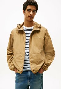 Chaqueta ligera color beige con capucha, que presenta puños elásticos y un cierre con cremallera. Llevada sobre una camiseta de rayas y vaqueros azules.