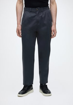 Mann trägt dunkelgraue Freizeithose mit Fronttaschen und schwarze Sneakers mit weißen Sohlen, steht vor einem schlichten Hintergrund.