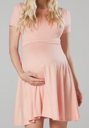 Femme enceinte aux longs cheveux blonds portant une robe rose clair à manches courtes, tenant son ventre d'une main.