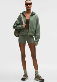 Groene cropped hoodie met ritssluiting, bruin hemdje, groene korte broek, grijze sneakers en zonnebril. De textuur is zacht, met atletische designelementen.