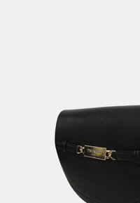 Borsa a spalla nera curvata in pelle, con una placca in metallo dorato con la scritta "TWINSET" e una tracolla sottile. Texture liscia, design minimal.