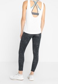 Top bianco senza maniche con scollatura rotonda, indossato sopra un reggiseno sportivo grigio. Leggings neri con un motivo testurizzato. Scarpe da ginnastica bianche.